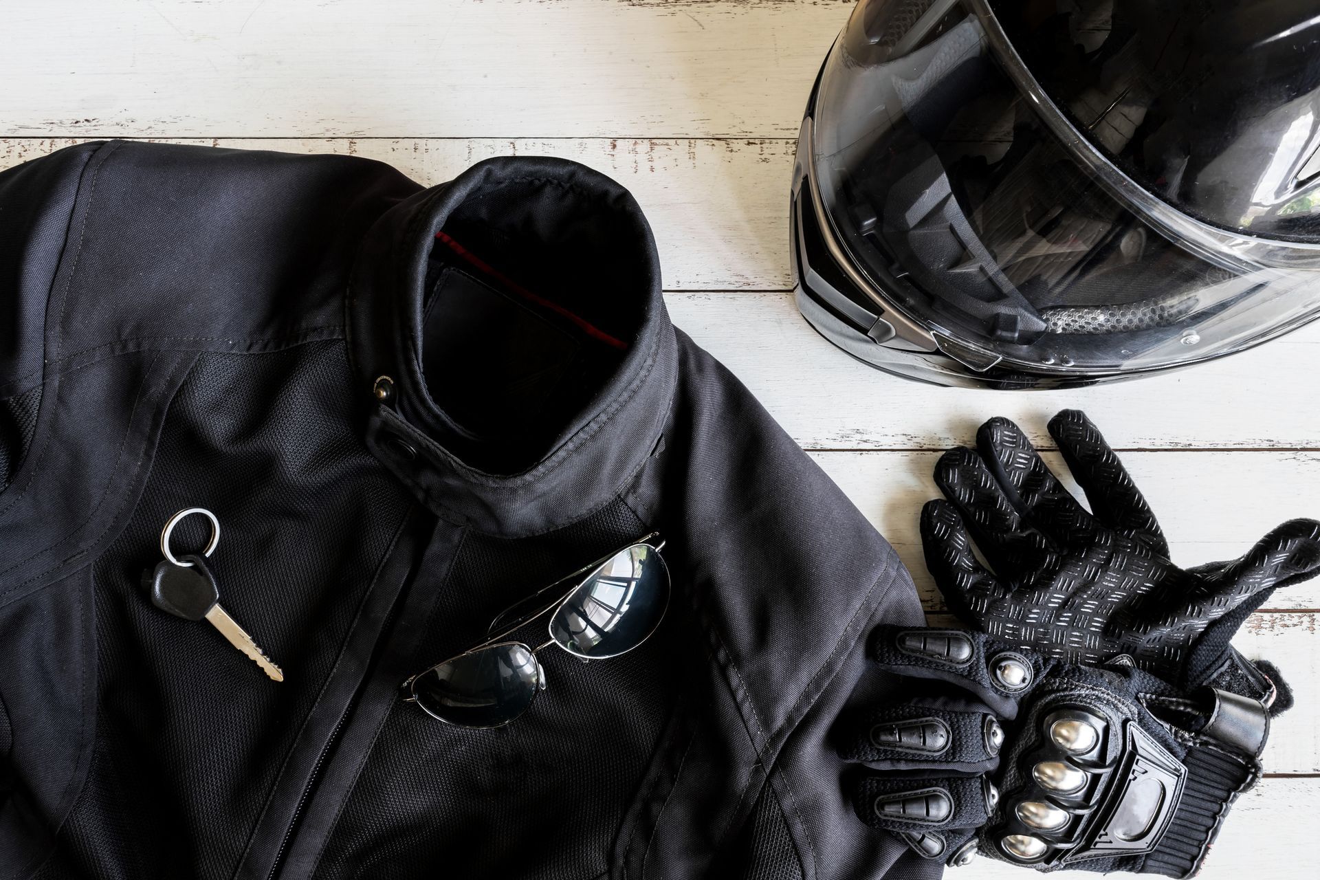 Black motorcycle jacket, helmet, gloves, sunglasses, and key on a white wooden surface.