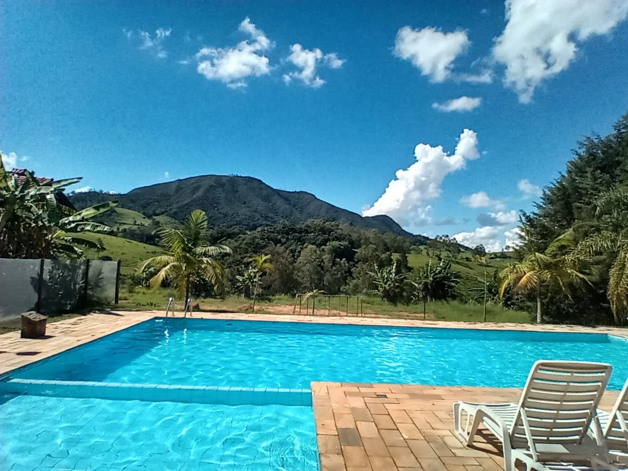 Piscina com vista para a montanha em um dia ensolarado. Água azul, nuvens brancas, árvores verdes.