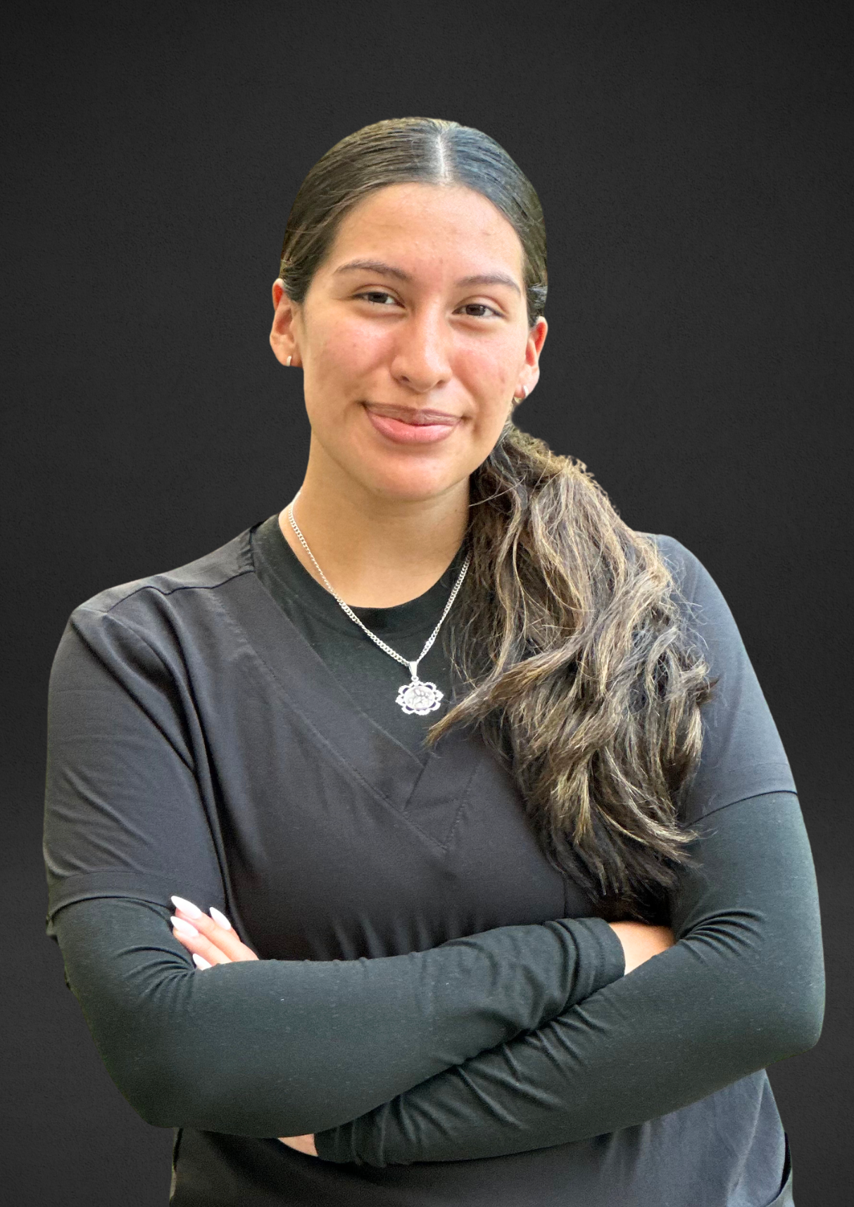 Woman with arms crossed, smiling, wearing black scrubs and a necklace. Against a dark background.