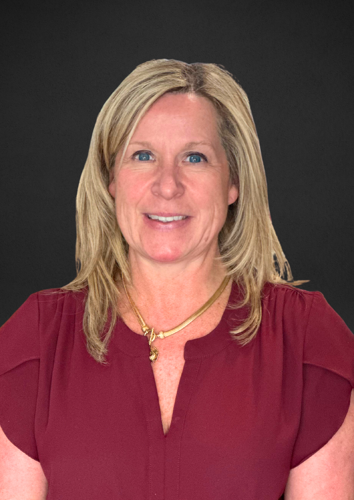 Woman with blonde hair wearing a maroon top and gold necklace smiling at the camera. Black background.