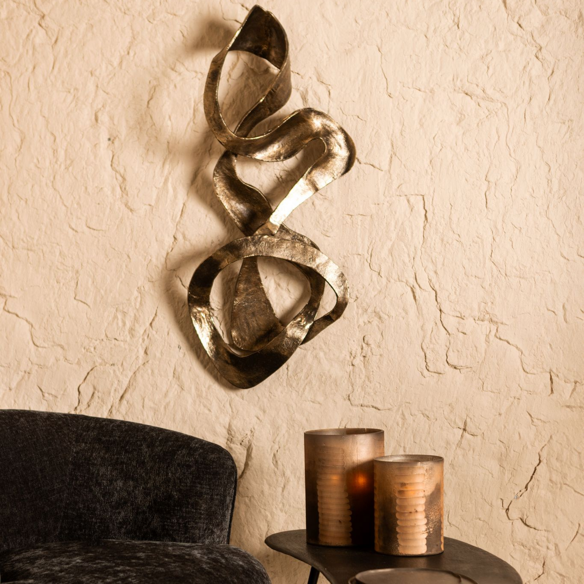 Abstracte metalen wanddecoratie boven een zwarte, pluizige stoel en een klein tafeltje met kaarsen.