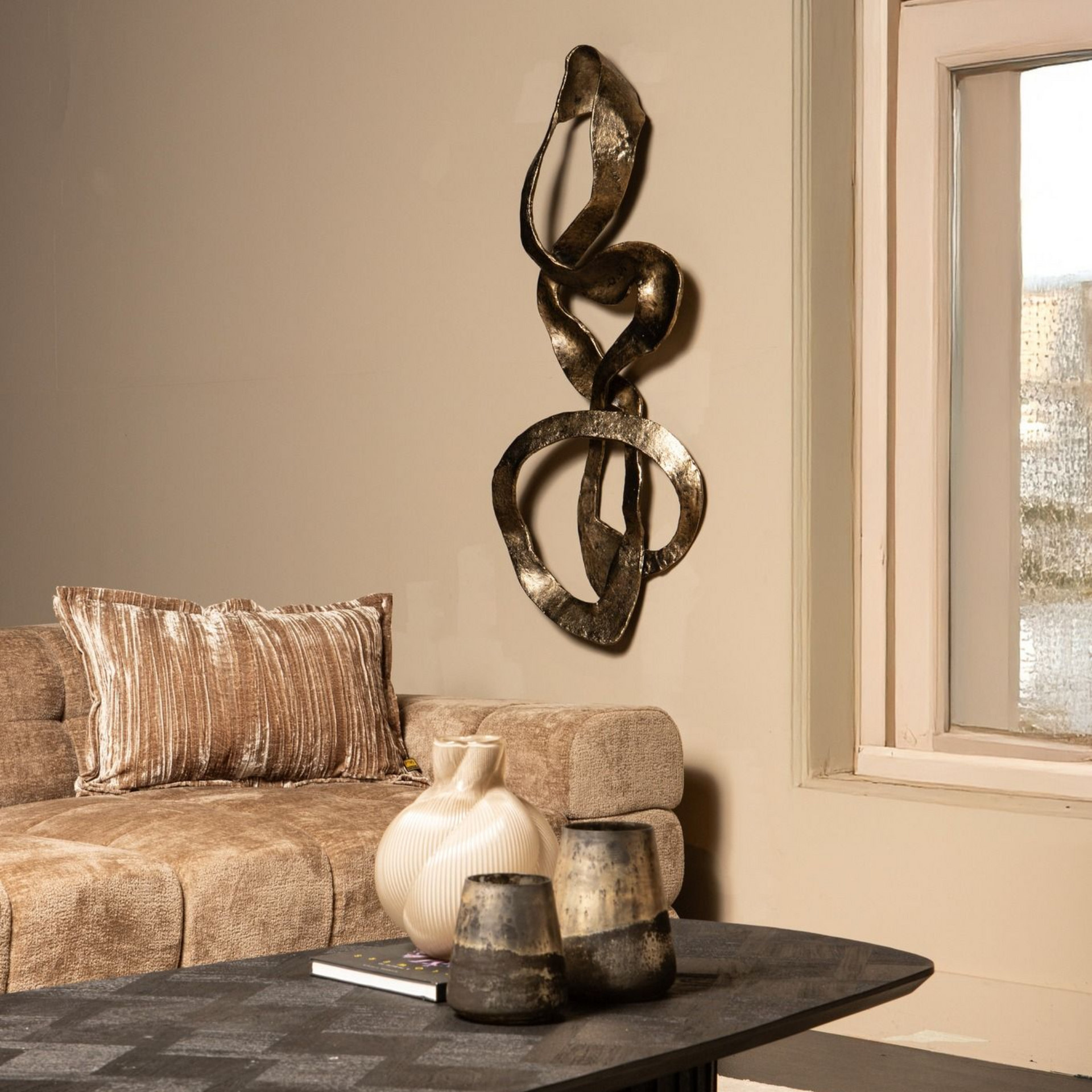Een eigentijdse, metalen wandsculptuur hangt boven een bank, met op de voorgrond een tafel met decoratieve vazen.
