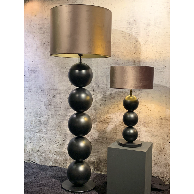 Staande en tafellampen, donkergrijze bollen op elkaar gestapeld, met bronzen trommelkappen, tegen een muur met structuur.
