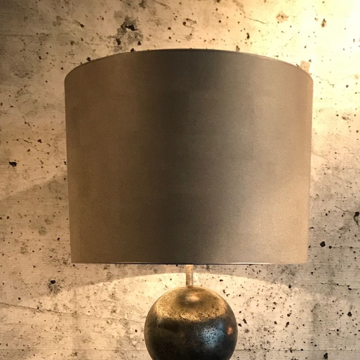 Zwarte lampenkap op een metallic gouden en zilveren lampvoet, tegen een grijze muur met structuur.