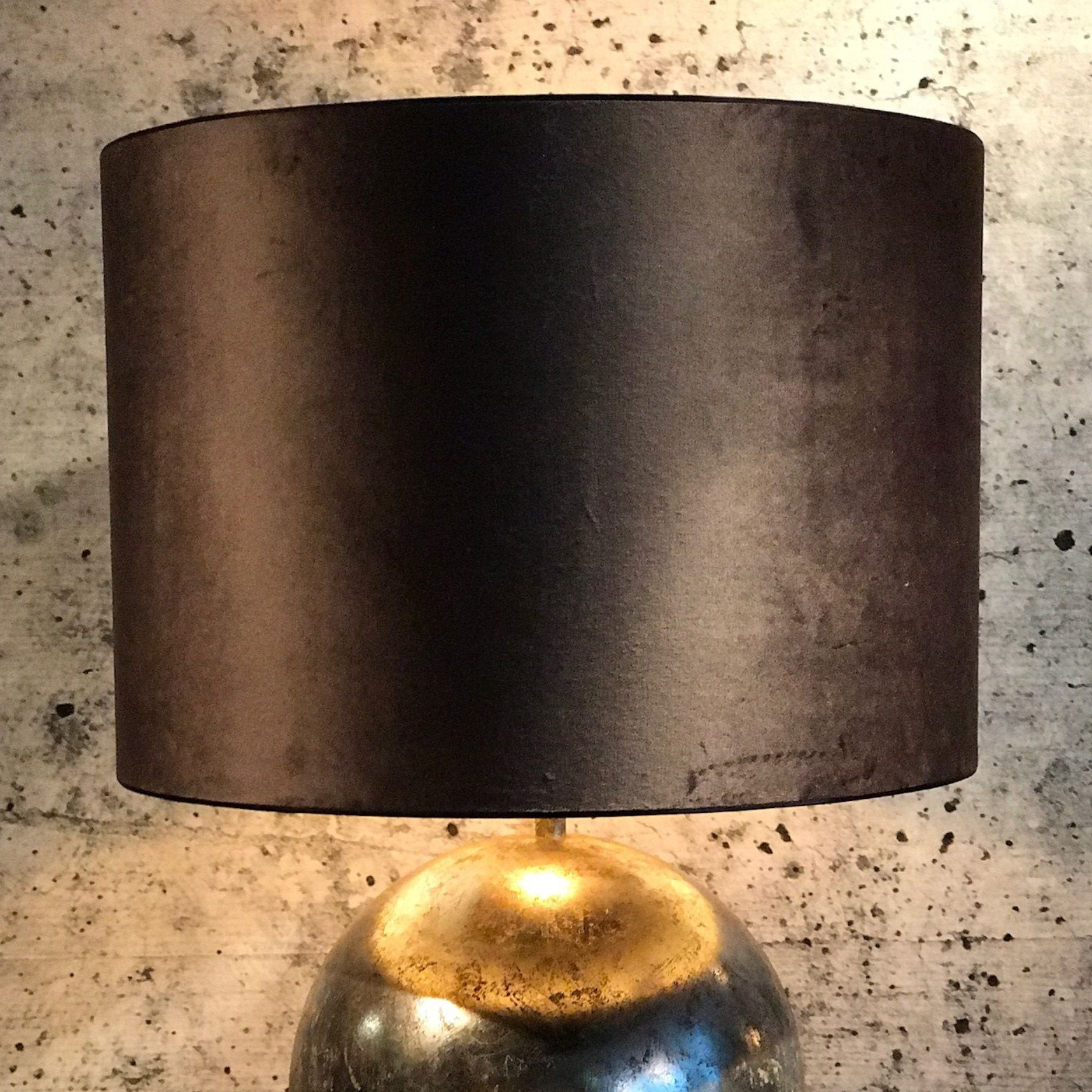 Zwarte lampenkap op een metallic gouden en zilveren lampvoet, tegen een grijze muur met structuur.
