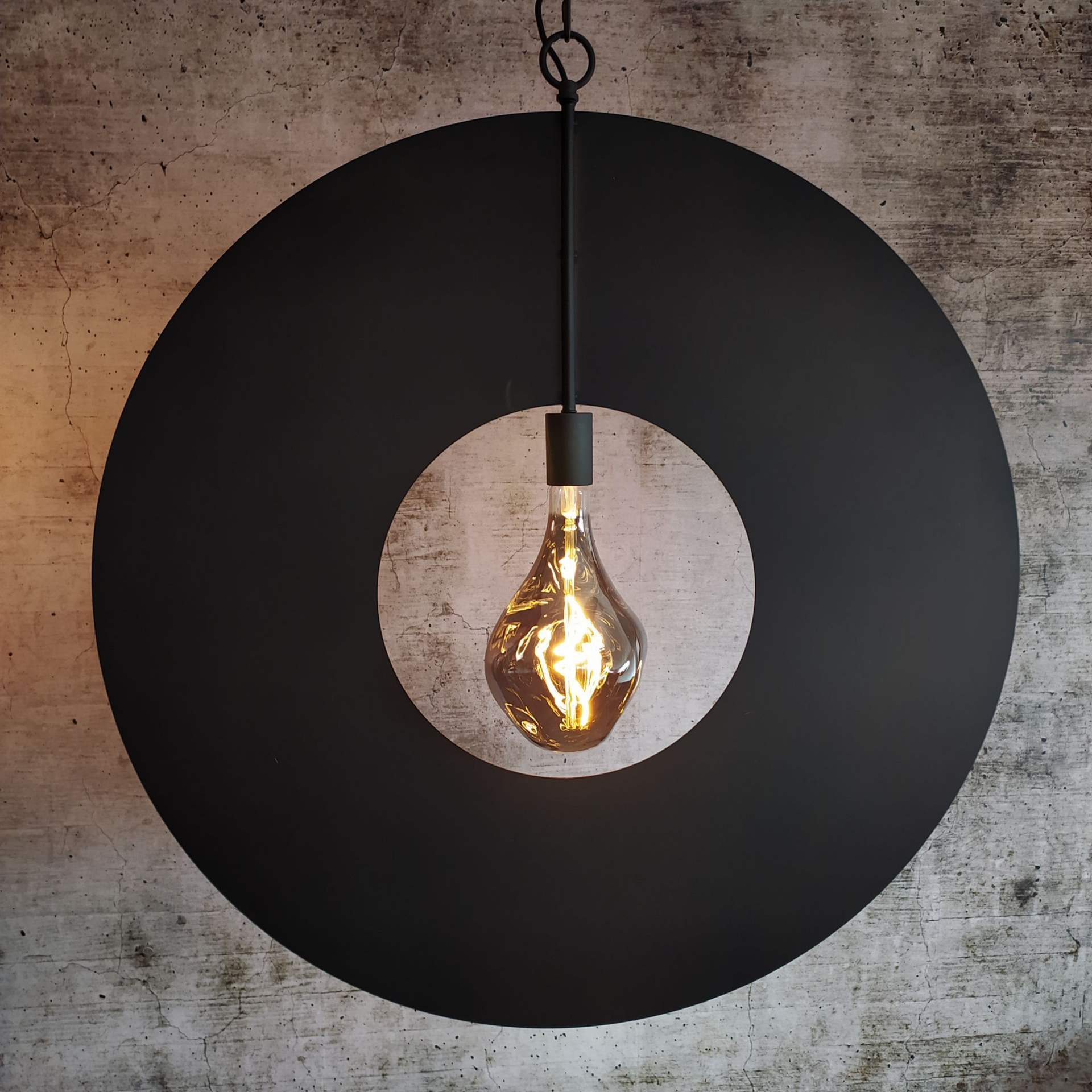 Zwarte ronde hanglamp met een grote lamp tegen een grijze muur met structuur.