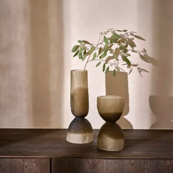 Twee matte keramische vazen ​​met bladerrijke takken staan ​​op een houten tafel tegen een beige muur.