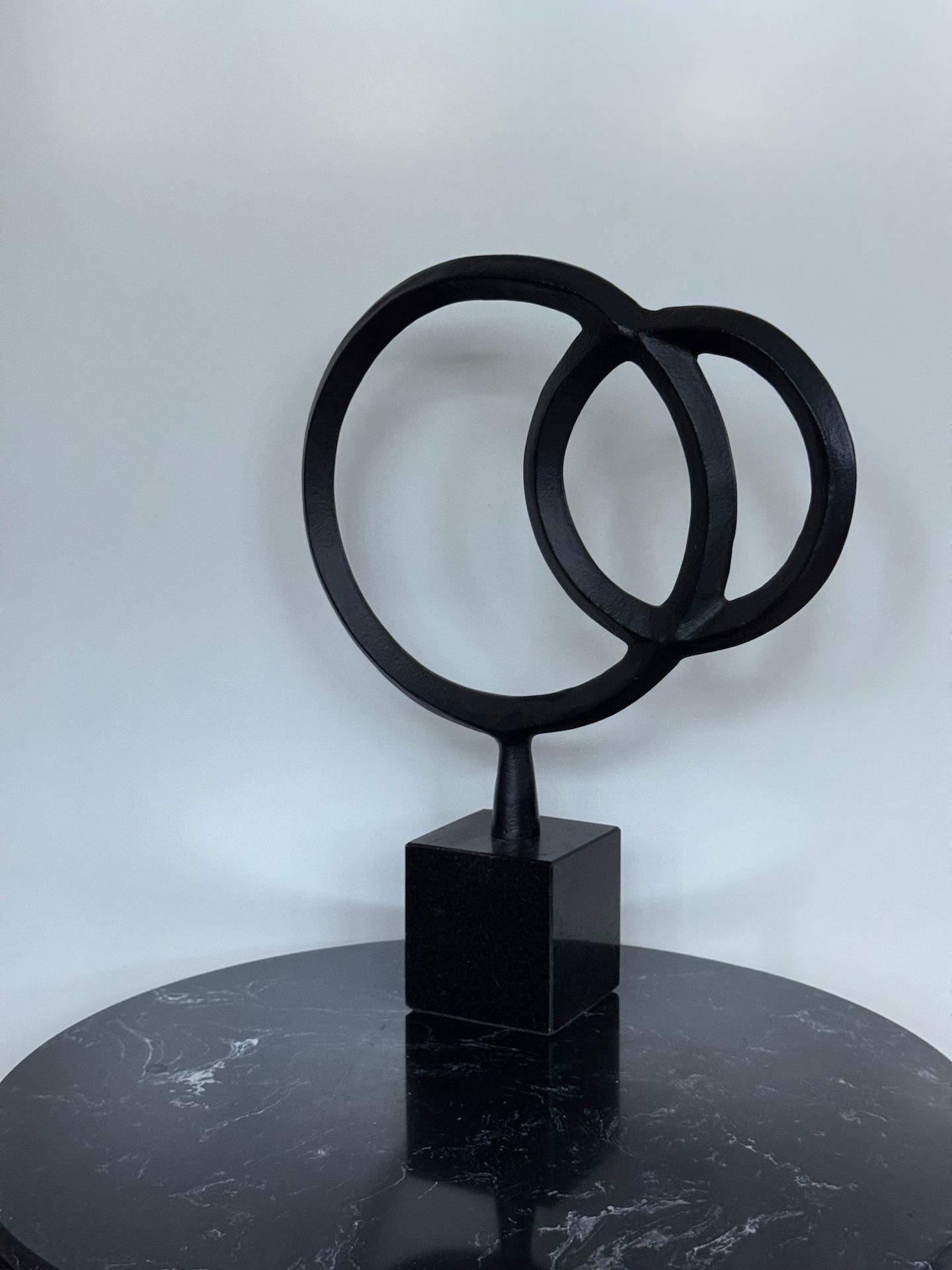 Een sculptuur van zwart metaal, bestaande uit twee elkaar kruisende ringen op een vierkante marmeren sokkel.