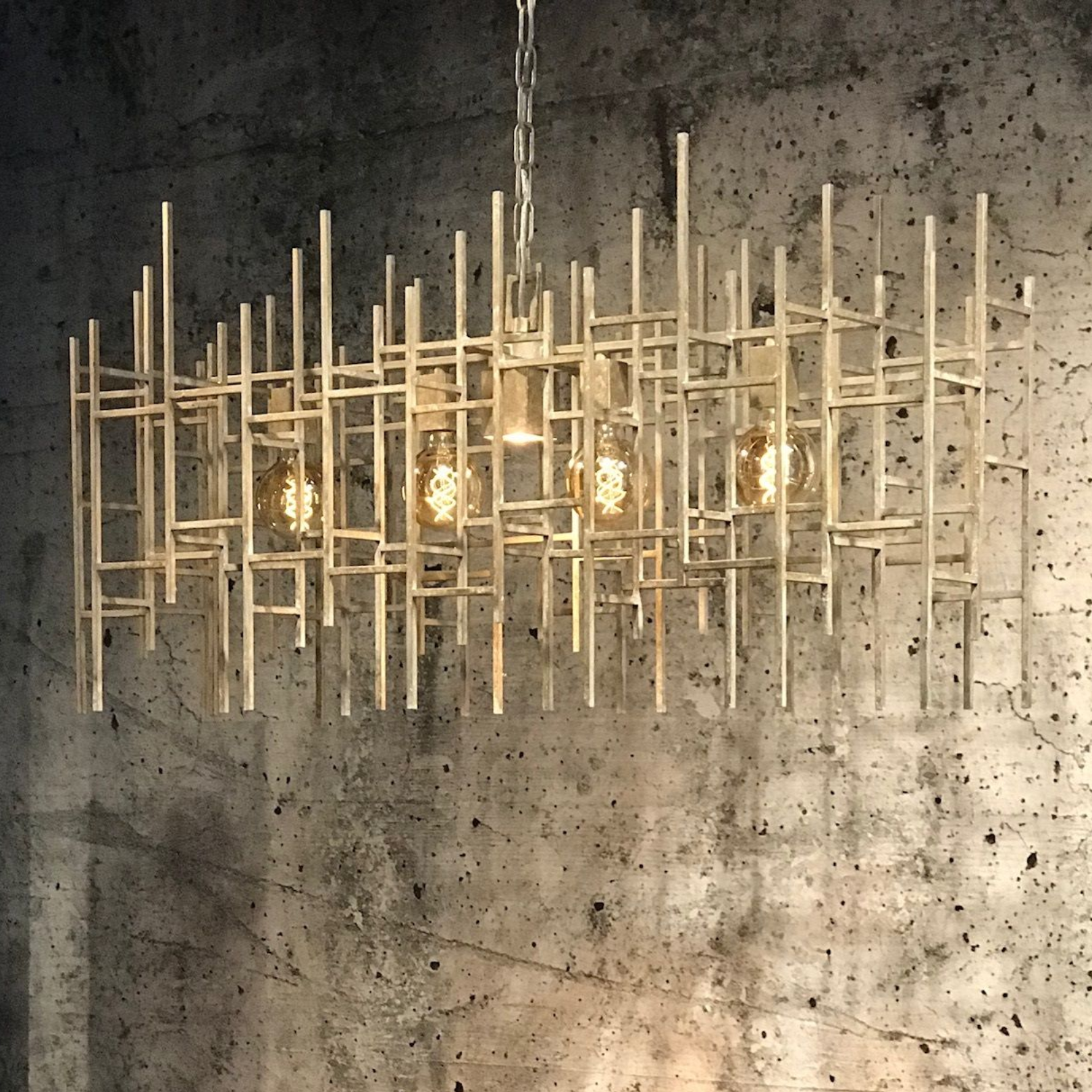Geometrische metalen kroonluchter met Edison-lampen, hangend tegen een muur van gestructureerd beton.