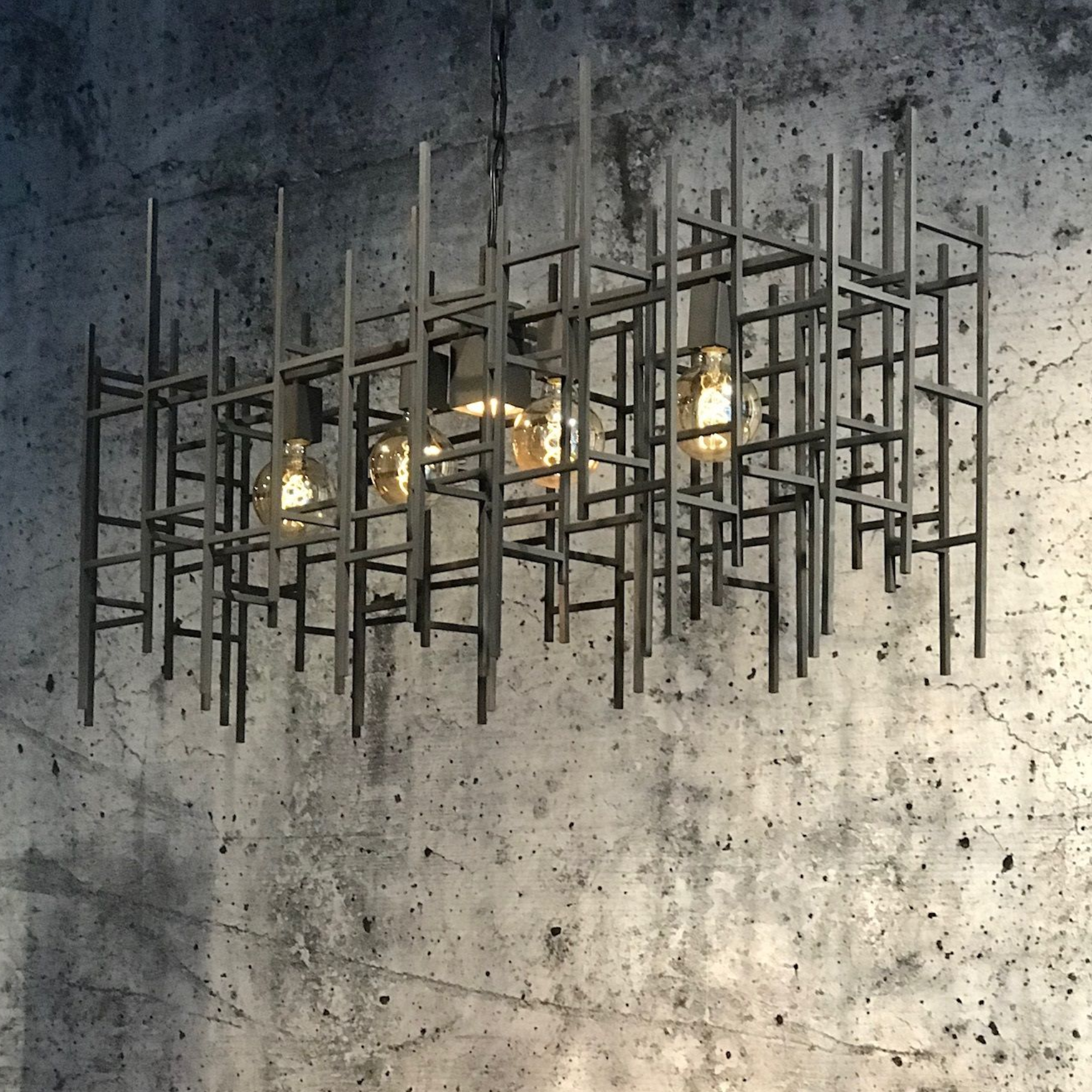 Geometrische metalen kroonluchter met Edison-lampen, hangend tegen een muur van gestructureerd beton.