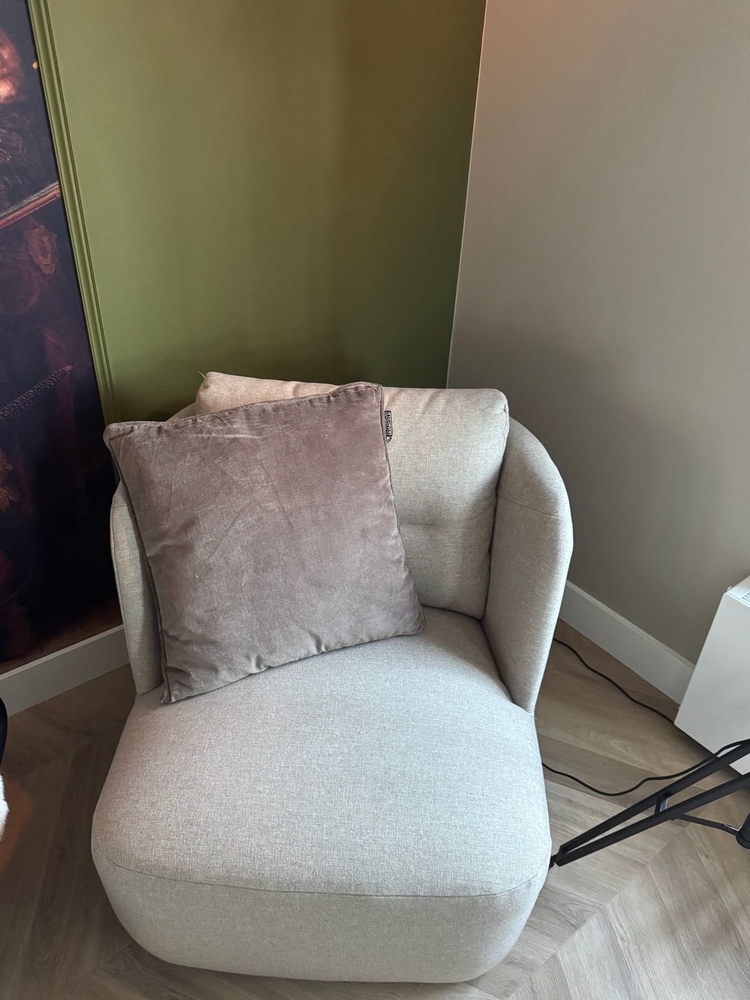 Een knusse, lichtgrijze fauteuil met een bruin kussen in een hoek van een kamer, met groene en grijze muren en een houten vloer.