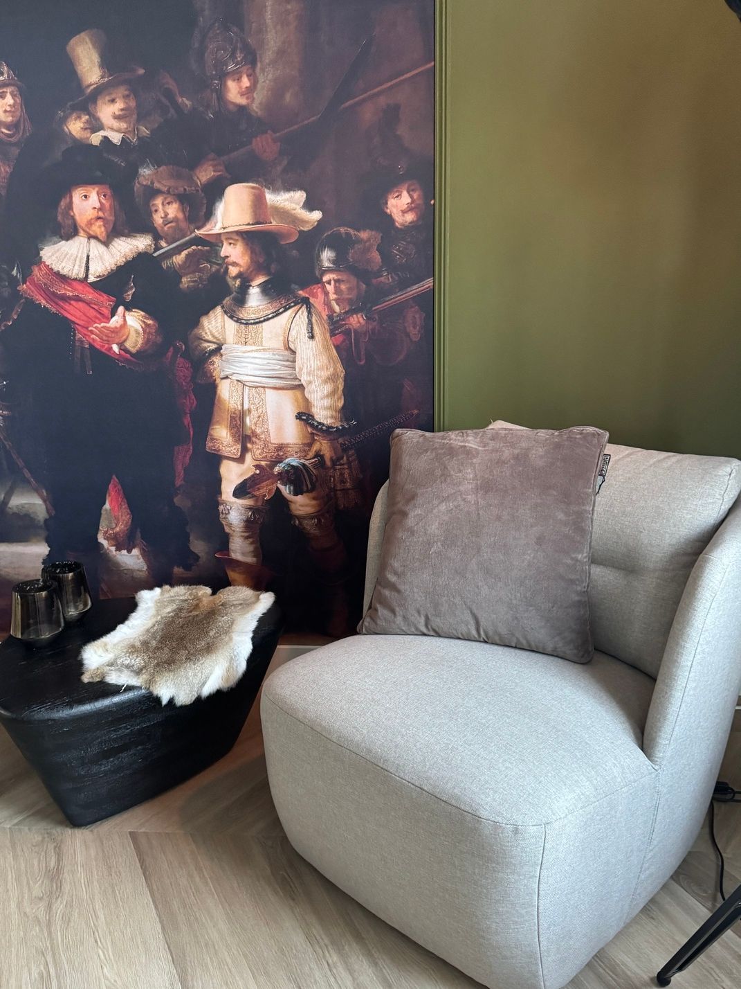 Een knusse, lichtgrijze fauteuil met een bruin kussen in een hoek van een kamer, met groene en grijze muren en een houten vloer.