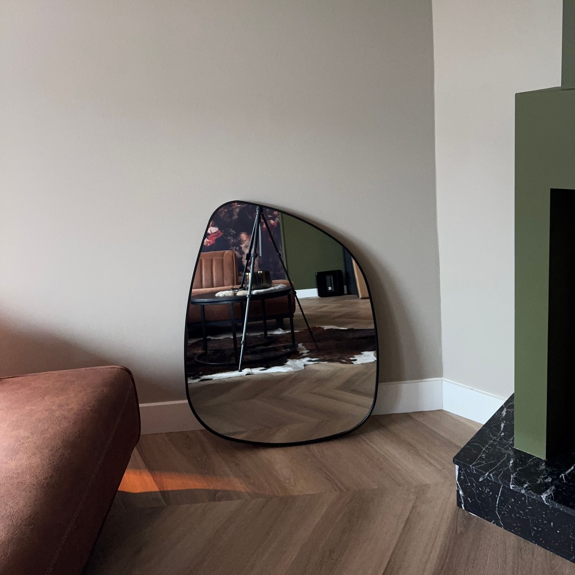 Een spiegel die tegen de muur leunt, weerspiegelt een woonkamer met een bruine bank, tafel en open haard.