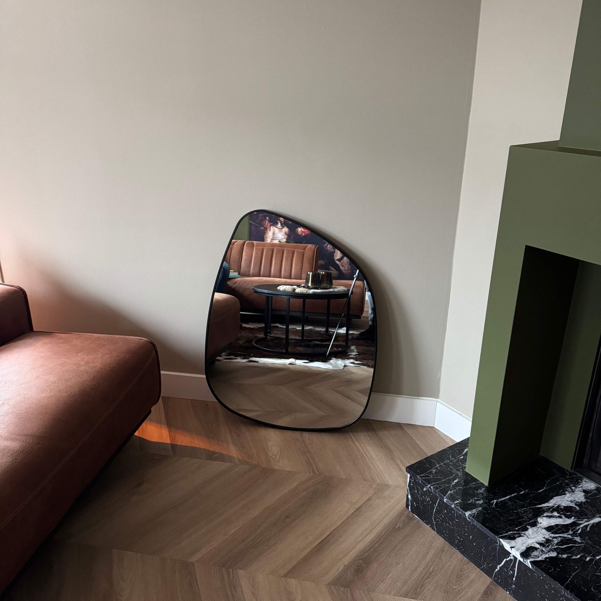 Een spiegel die tegen de muur leunt, weerspiegelt een woonkamer met een bruine bank, tafel en open haard.