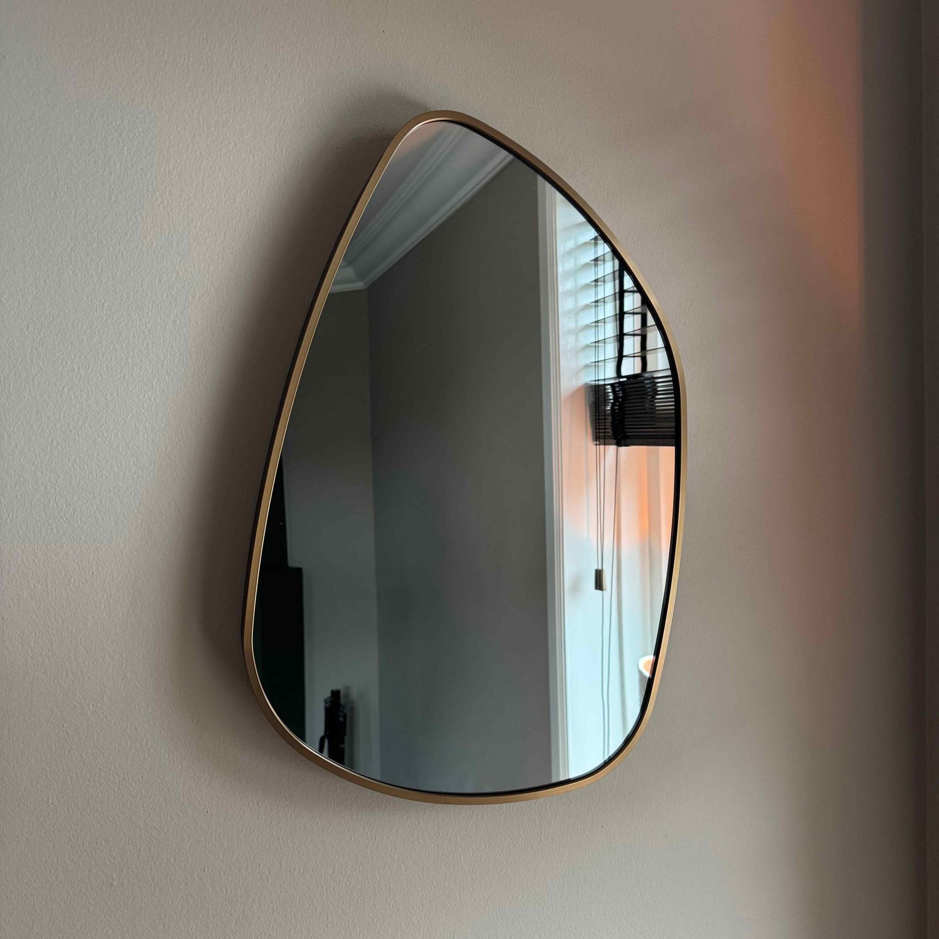 Een kiezelvormige spiegel met een gouden frame hangt aan een beige muur en weerspiegelt een raam met jaloezieën.