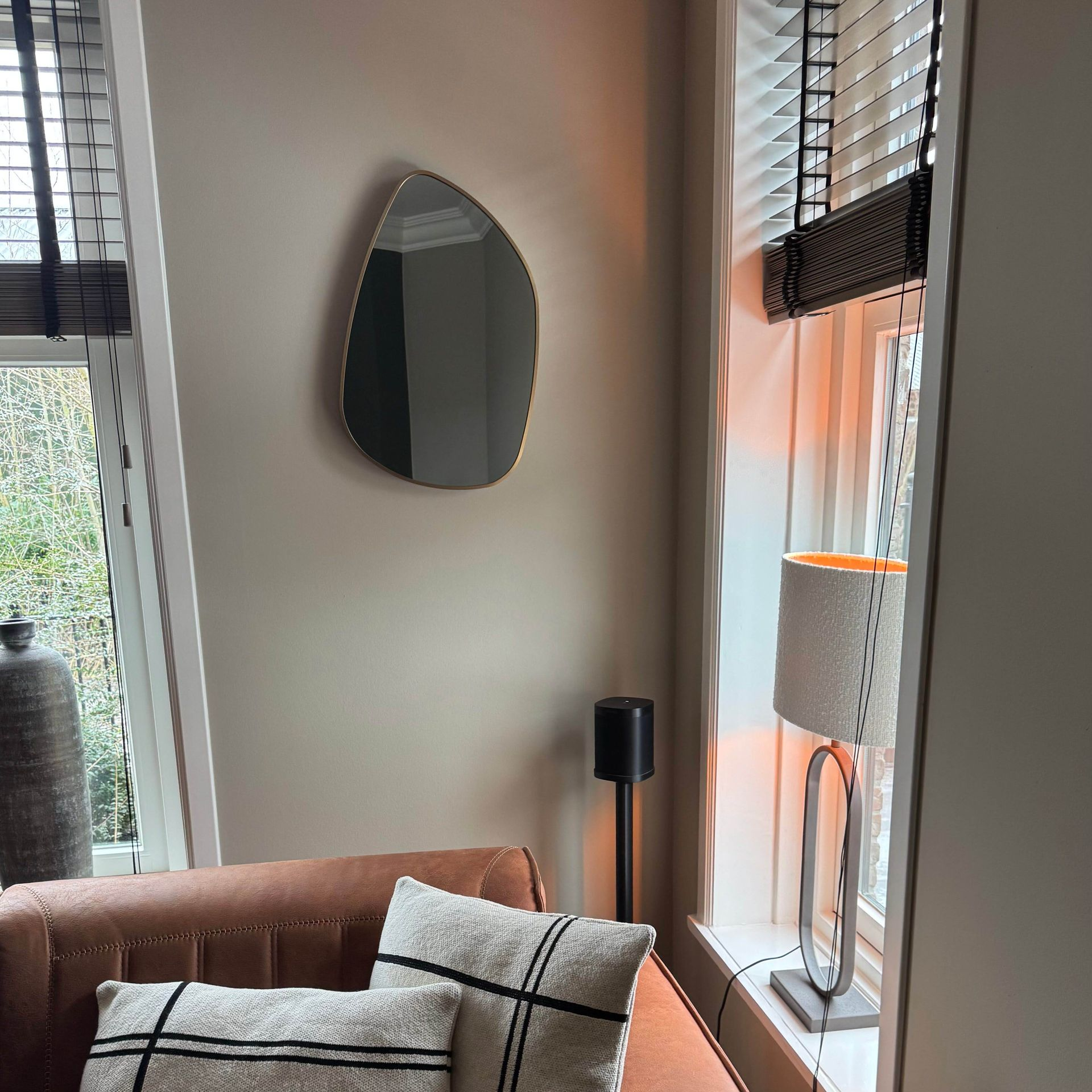 Een kiezelvormige spiegel met een gouden frame hangt aan een beige muur en weerspiegelt een raam met jaloezieën.