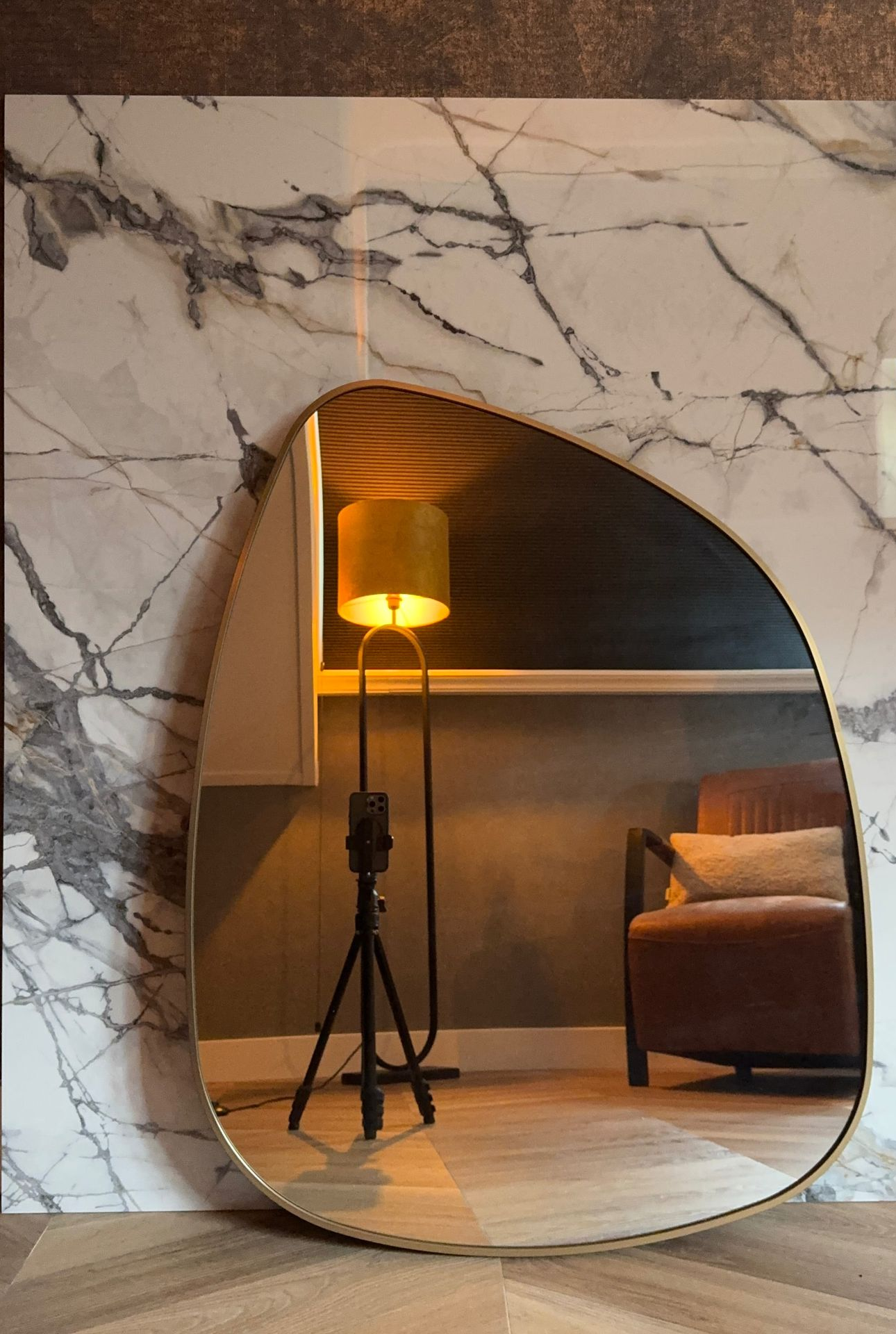 Een spiegel die een kamer met een lamp en een fauteuil weerspiegelt, tegen een marmeren muur.