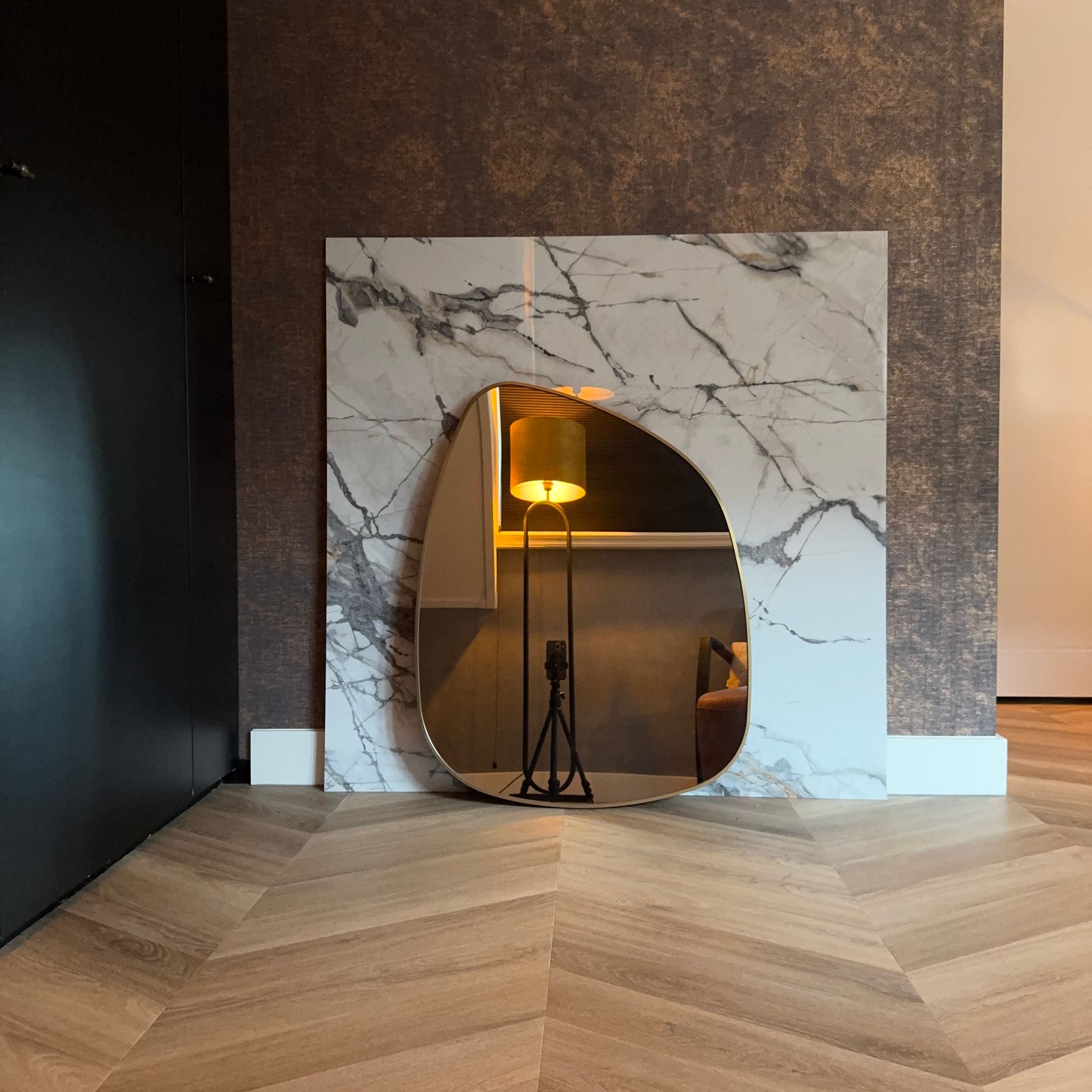 Een spiegel die een kamer met een lamp en een fauteuil weerspiegelt, tegen een marmeren muur.