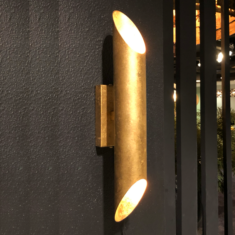 Goudkleurige, cilindrische wandlamp op een donkergrijze muur, die zowel naar boven als naar beneden schijnt.