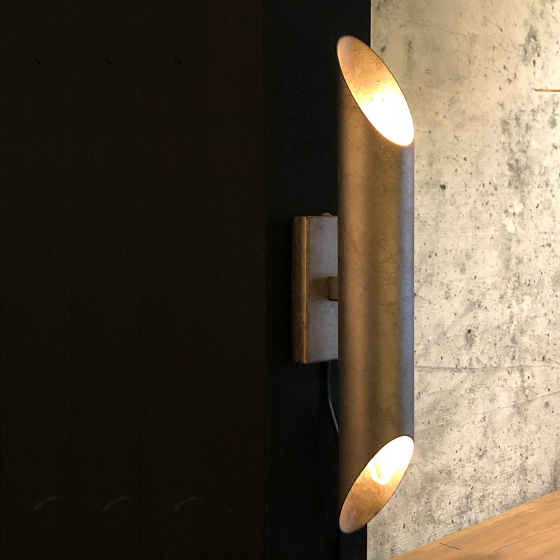 Goudkleurige, cilindrische wandlamp op een donkergrijze muur, die zowel naar boven als naar beneden schijnt.