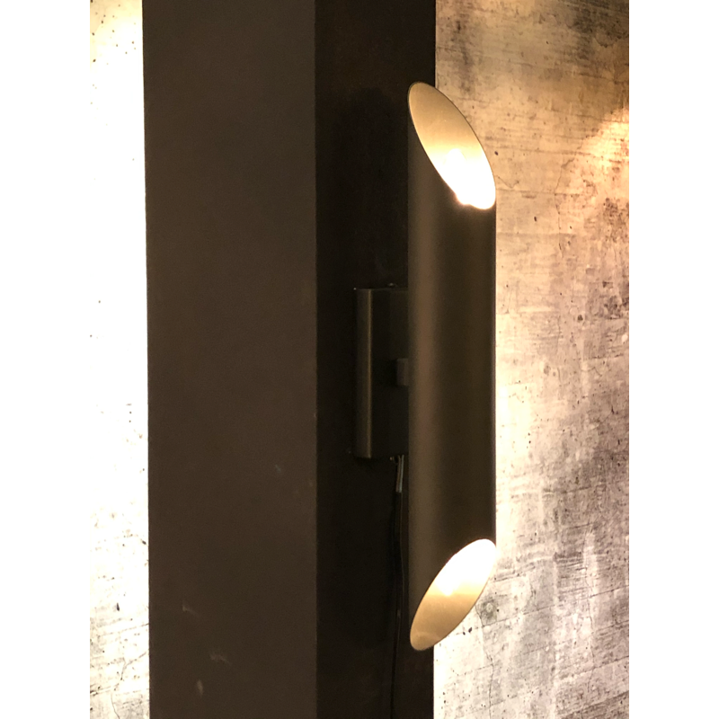 Donkere, cilindrische wandlamp met lichtuitstraling aan beide uiteinden; gemonteerd op een donkere pilaar en betonnen muur.