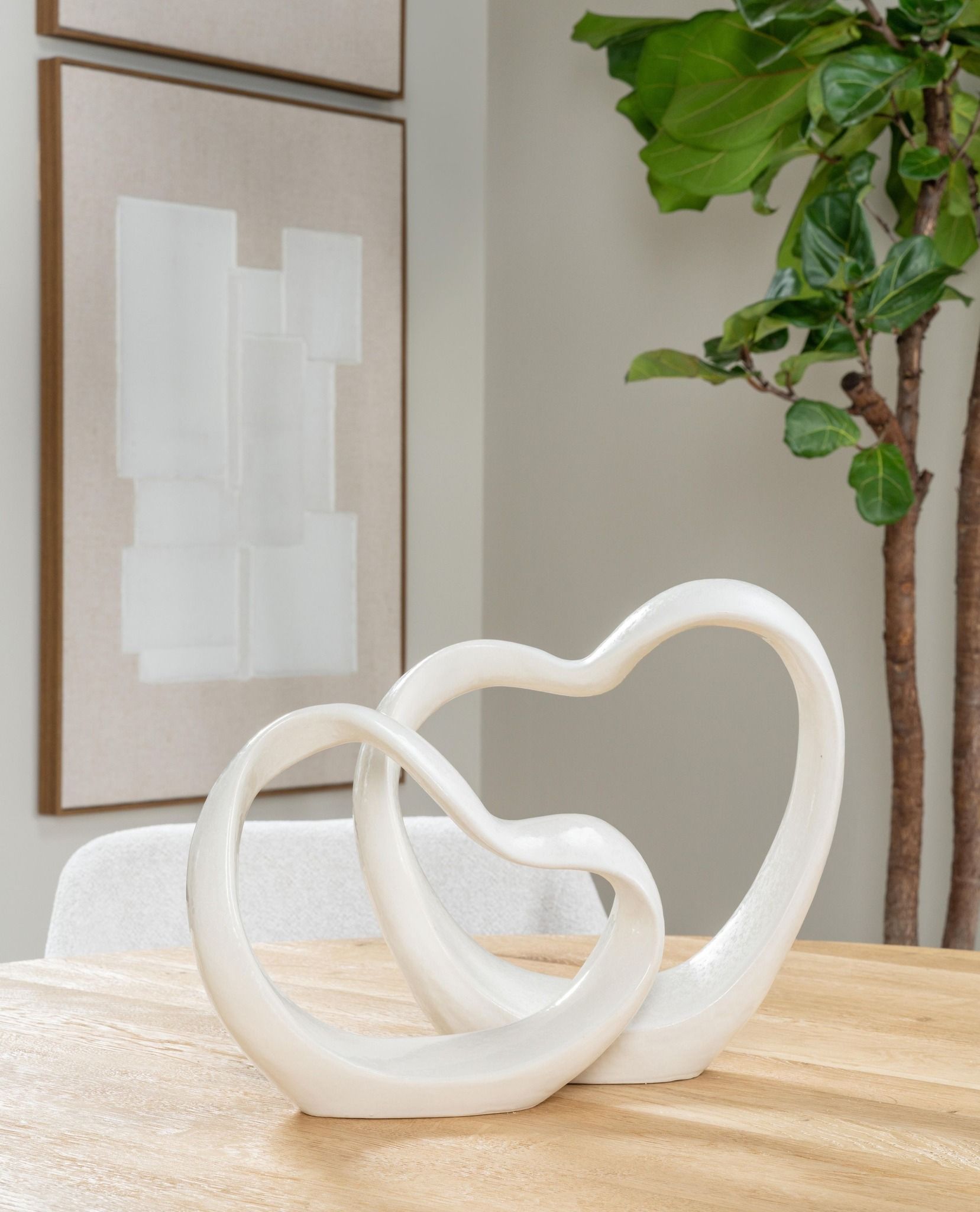 Een wit, hartvormig beeldje op een lichte houten tafel, met abstracte kunst en een plant op de achtergrond.