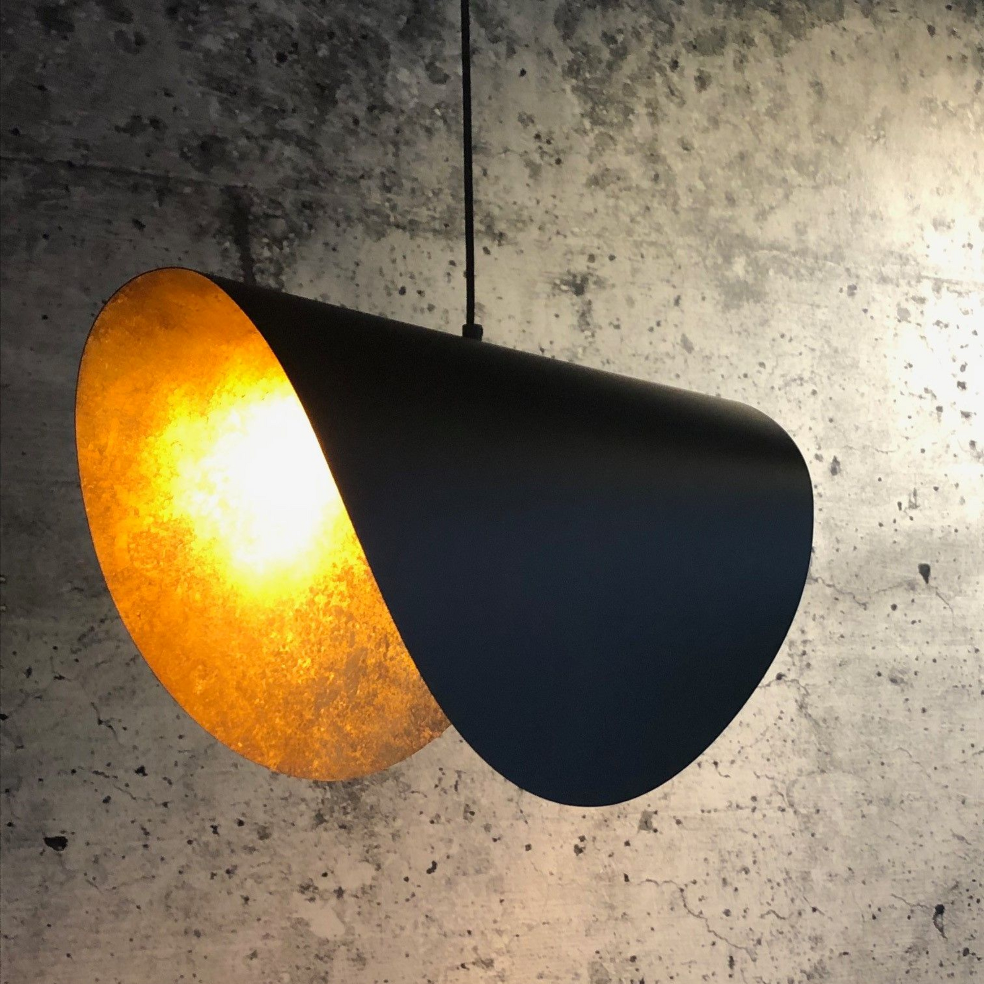 Zwarte hanglamp met gouden binnenkant, hangend tegen een grijze muur met structuur.