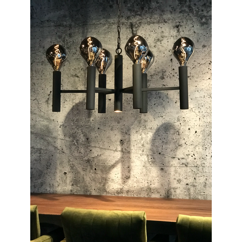 Een zwarte kroonluchter met grote gloeilampen hangt boven een houten tafel met groene stoelen.
