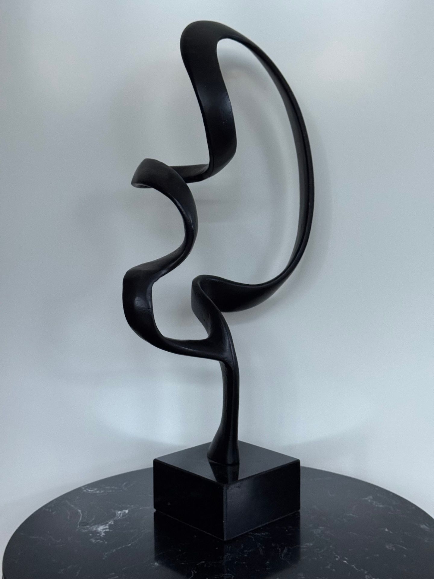 Abstracte sculptuur van zwart metaal op een zwarte vierkante sokkel, die in een lus naar boven buigt.