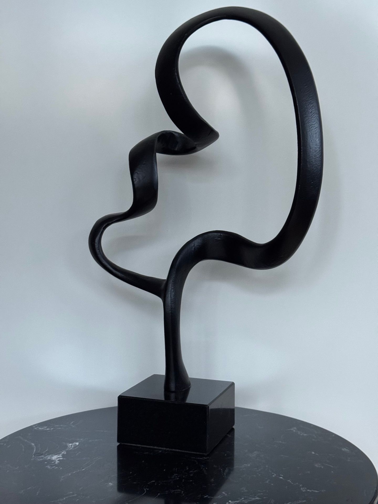 Abstracte sculptuur van zwart metaal op een zwarte vierkante sokkel, die in een lus naar boven buigt.