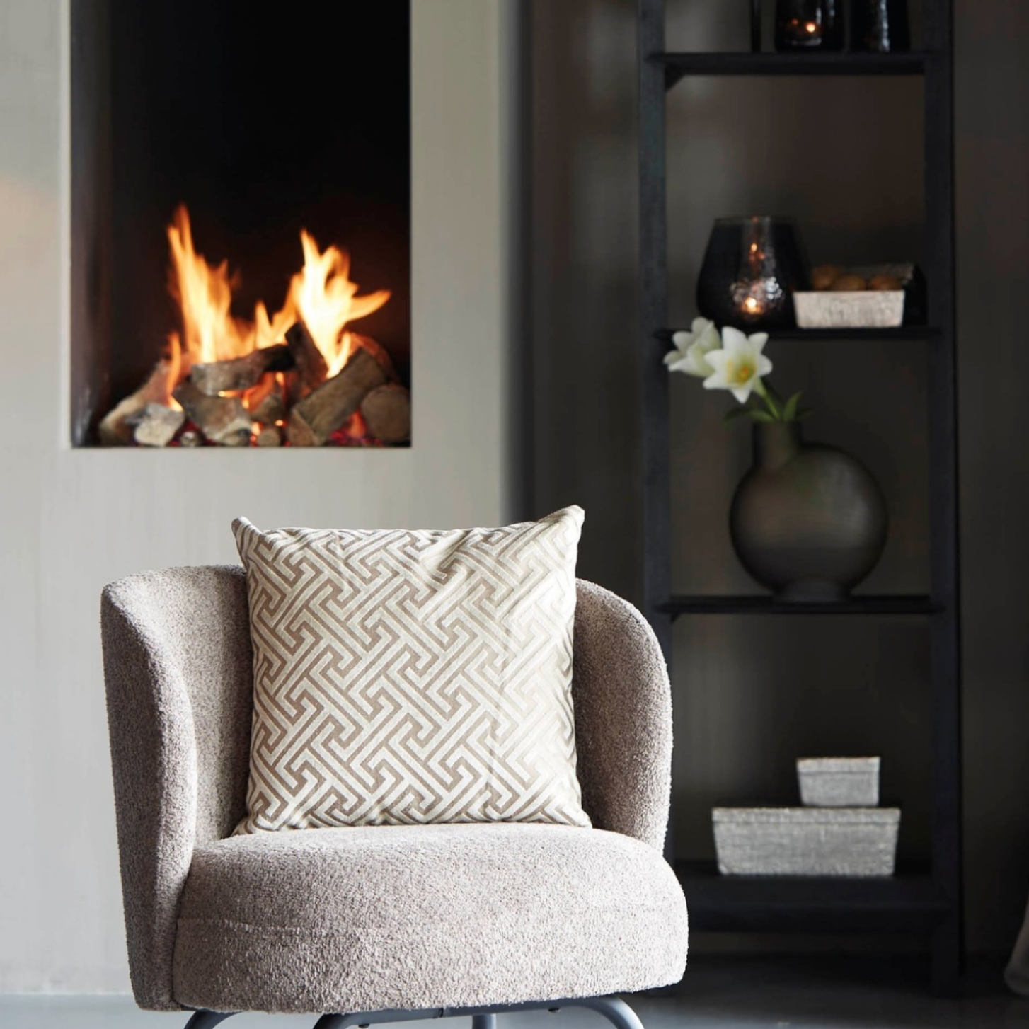 Gezellige woonkamer met een brandende open haard, een sierkussen op een stoel en een boekenkast vol decoratie.