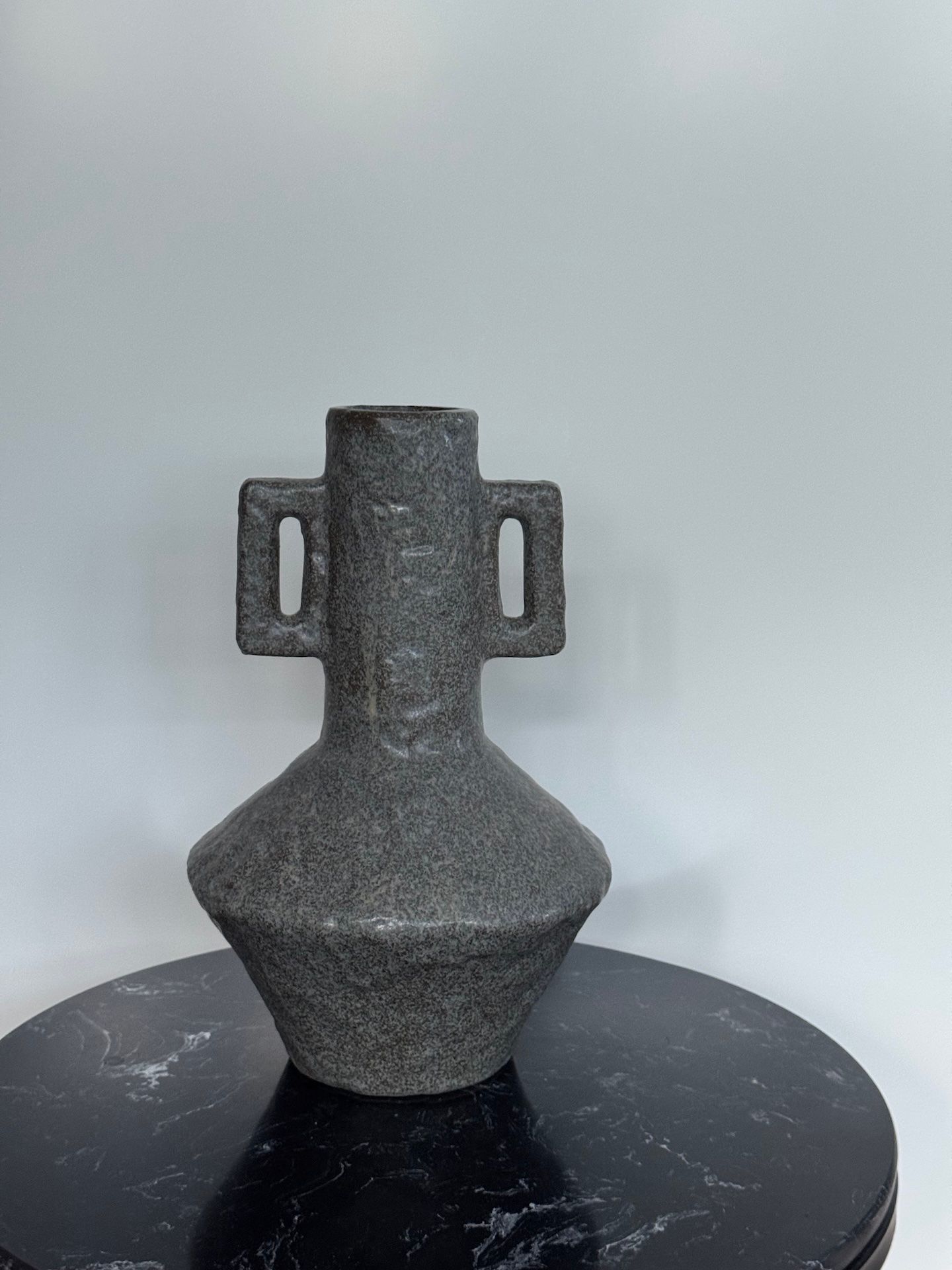 Donkere, gestructureerde vaas van keramiek met handvatten, staand op een zwart, gemarmerd oppervlak.