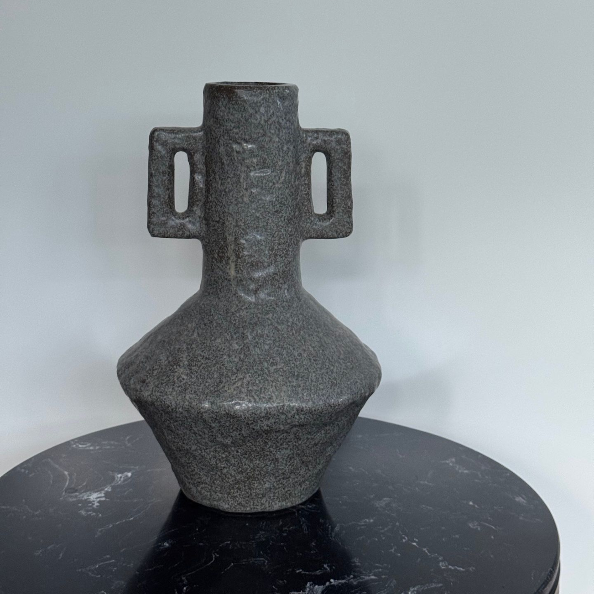 Donkere, gestructureerde vaas van keramiek met handvatten, staand op een zwart, gemarmerd oppervlak.