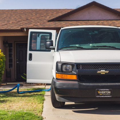 White van parked in front of a house, door open, hose running, sunlit daytime scene.