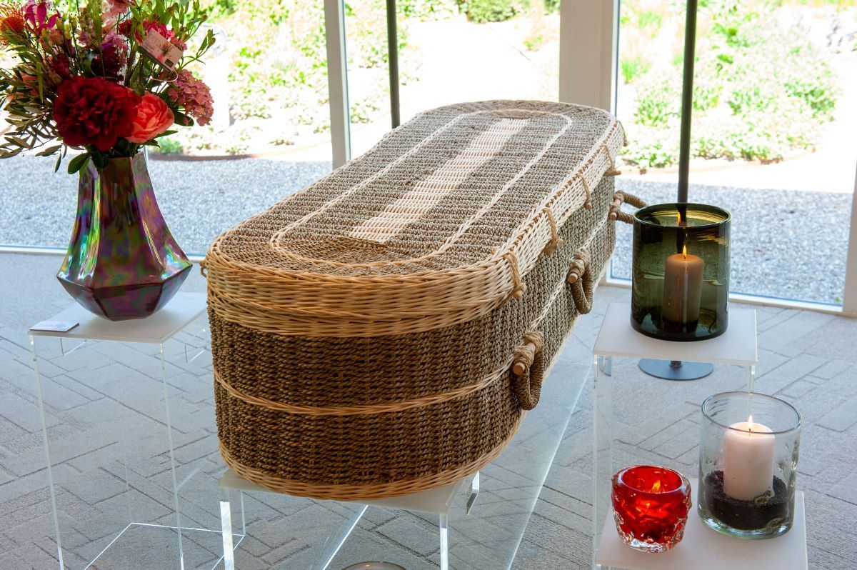 Wicker casket on a glass table, with flowers and candles, in a bright room with windows.