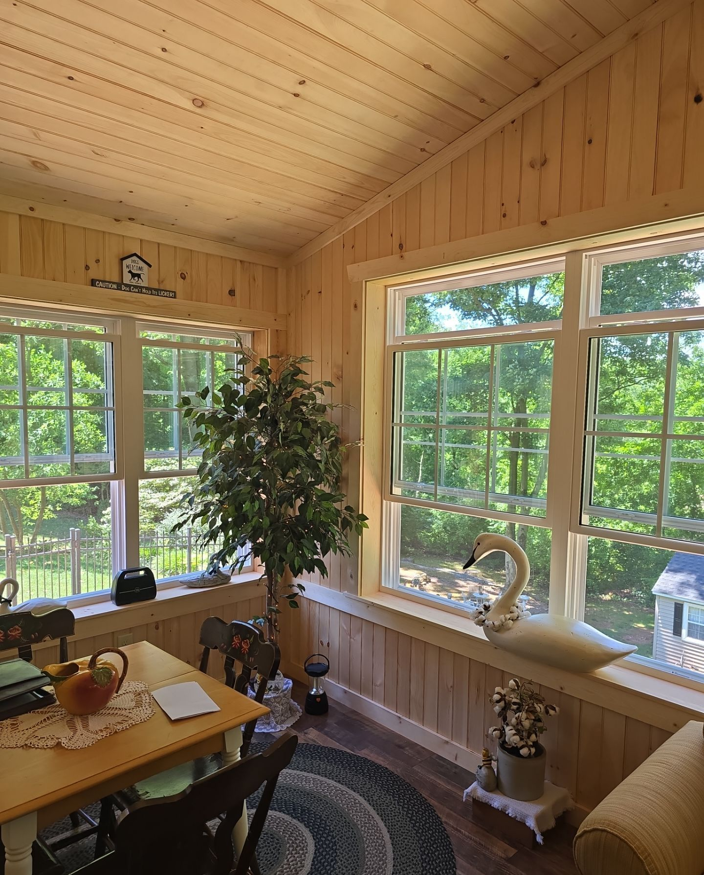 Sunroom with white sofa and ottoman, surrounded by large windows overlooking a yard.