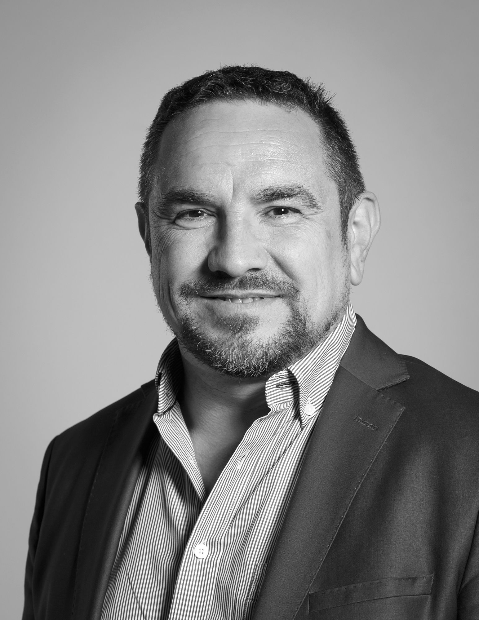 Un homme avec une barbe porte un costume et sourit sur une photo en noir et blanc.