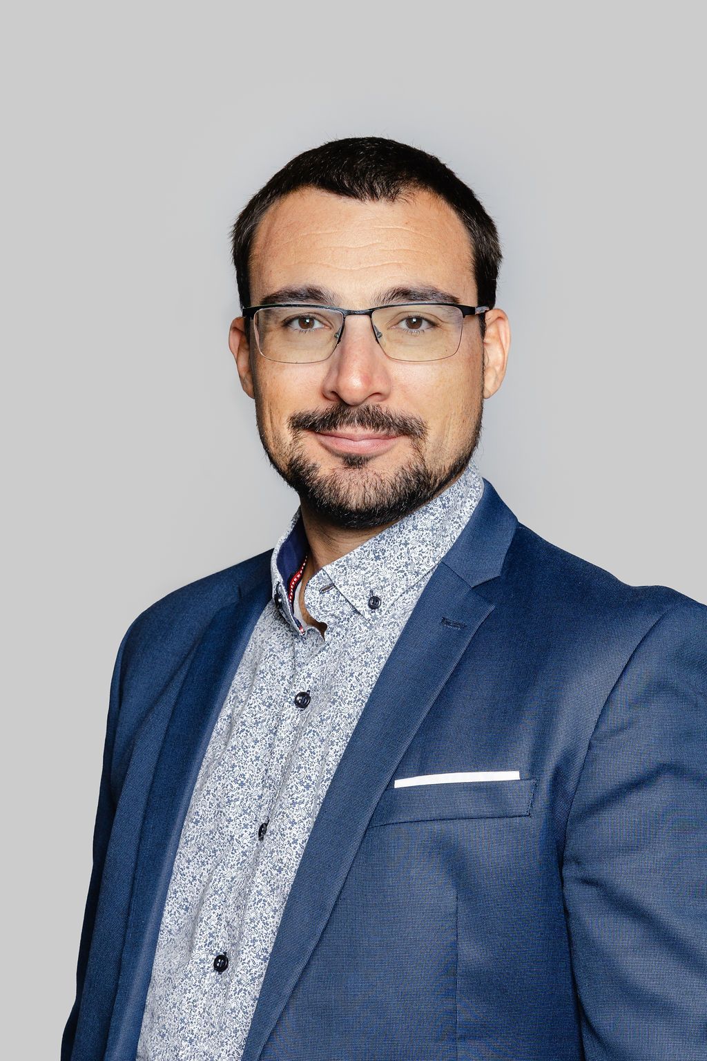 Homme à lunettes, blazer bleu, chemise à motifs, souriant, fond neutre.