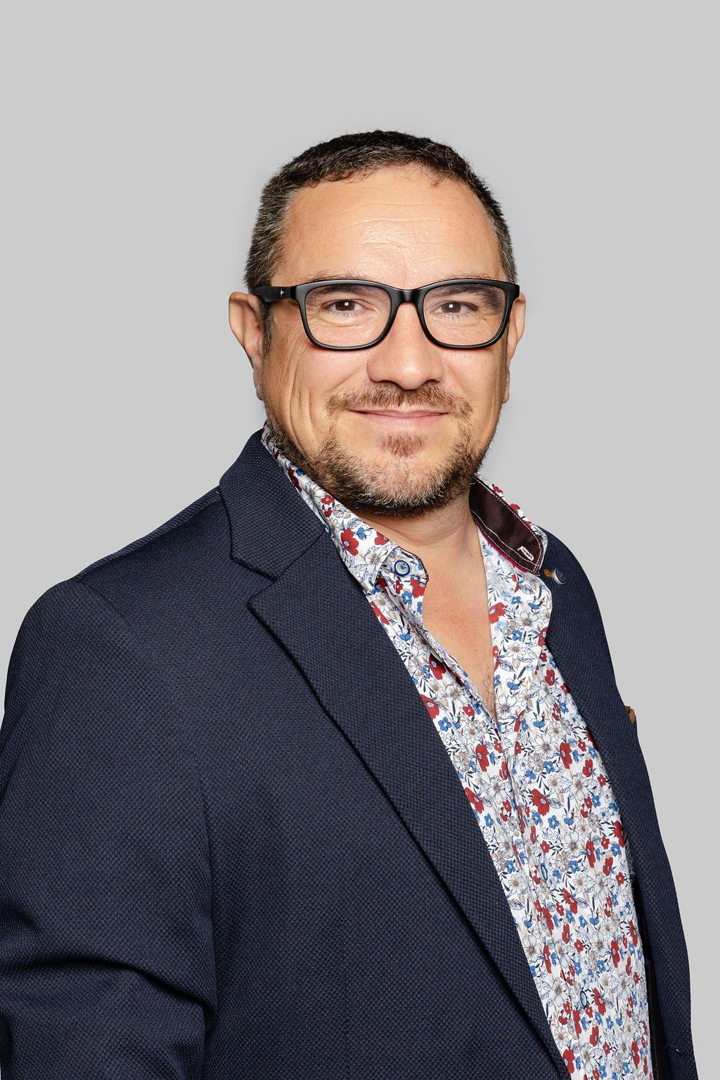 Un homme en blazer et chemise à motifs sourit à l'objectif. Le portrait est en noir et blanc.