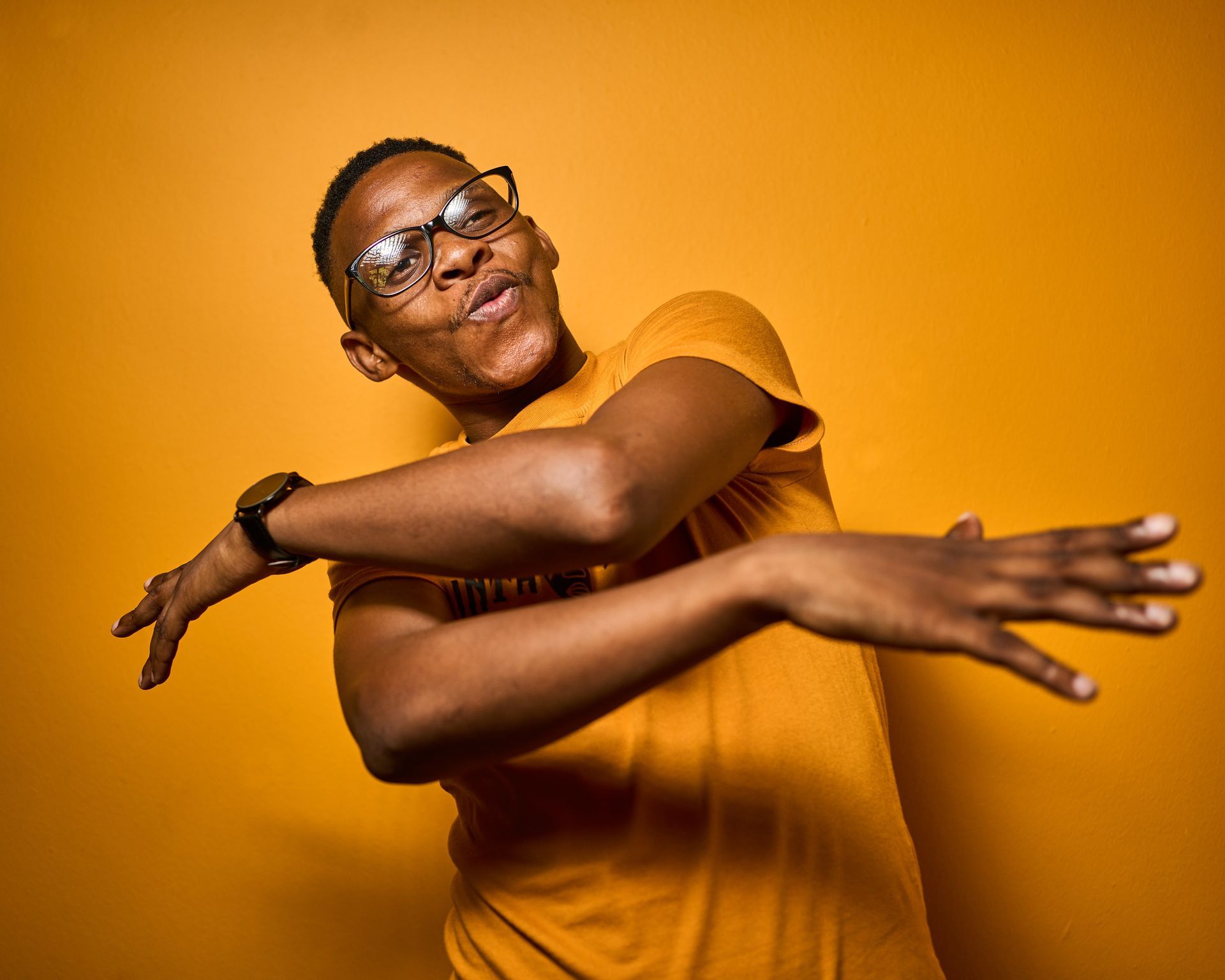 A man wearing glasses and a yellow shirt is dancing in front of a yellow background.