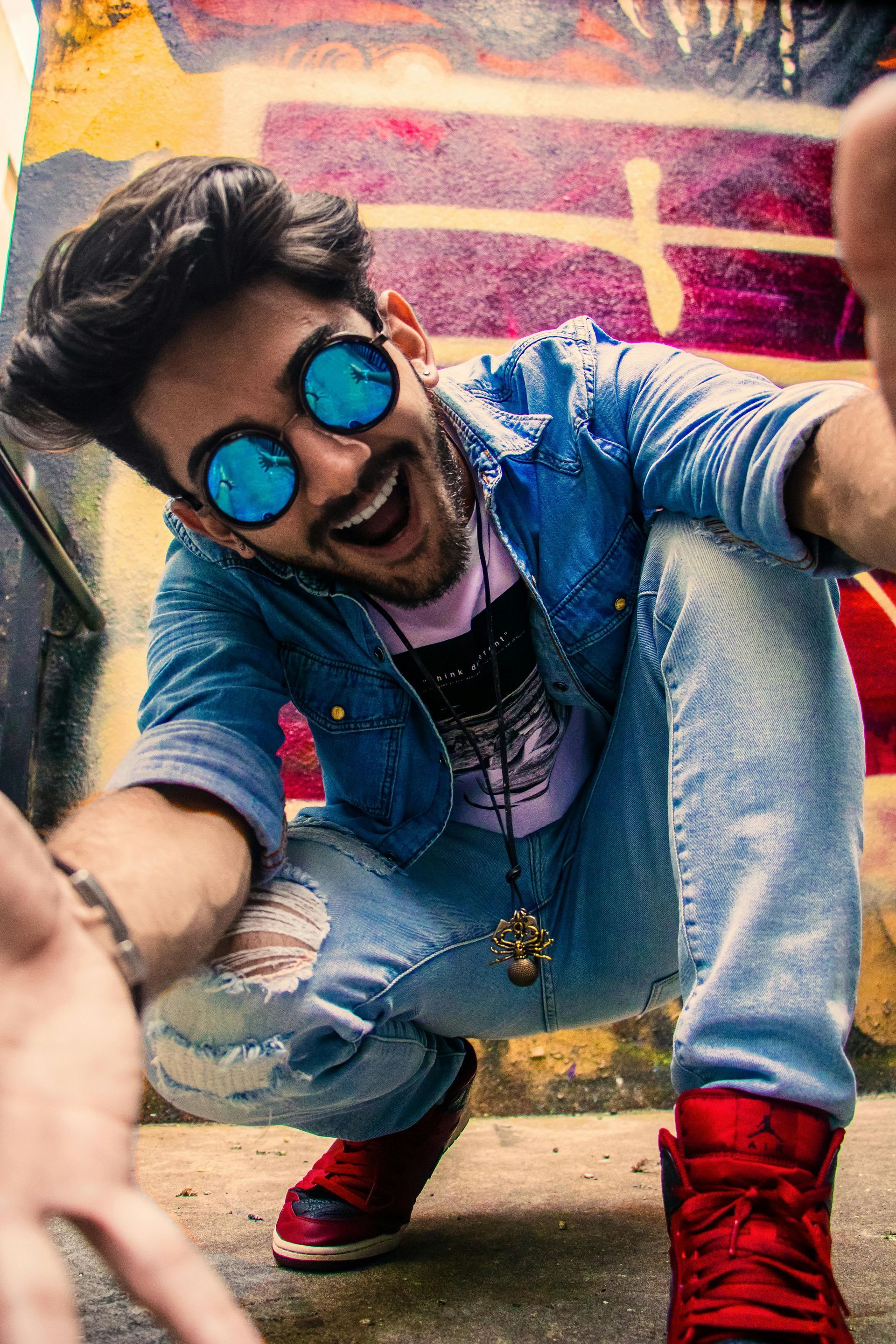 A man wearing sunglasses is kneeling down in front of a graffiti wall.