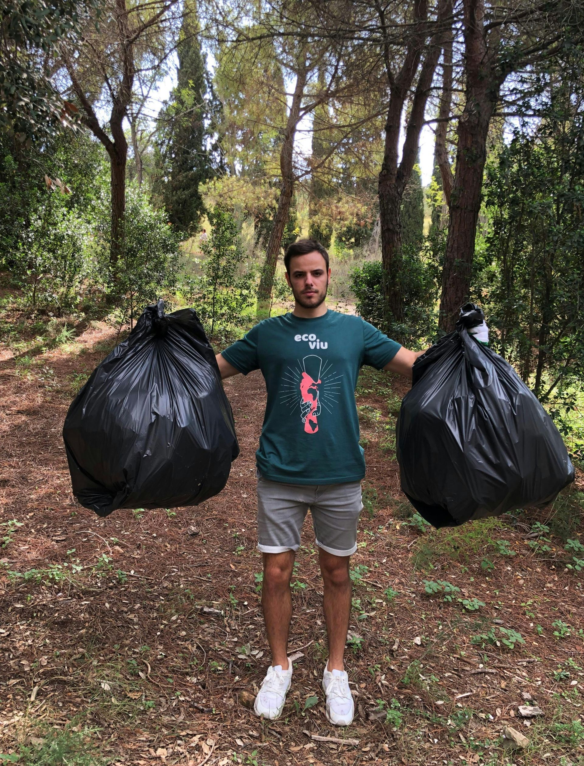 A man is holding two trash bags in his hands in the woods.