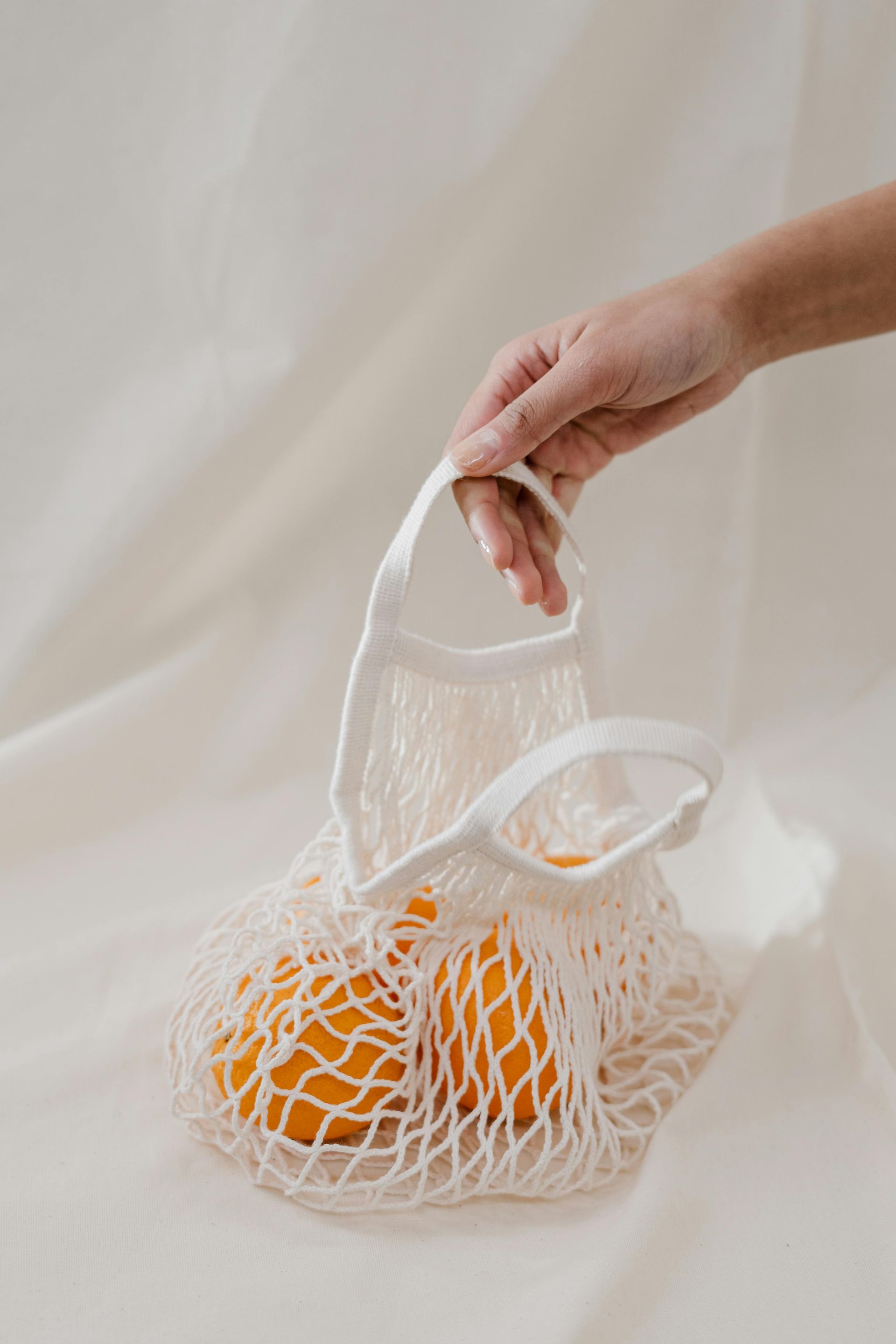 A person is holding a white net bag with oranges in it.