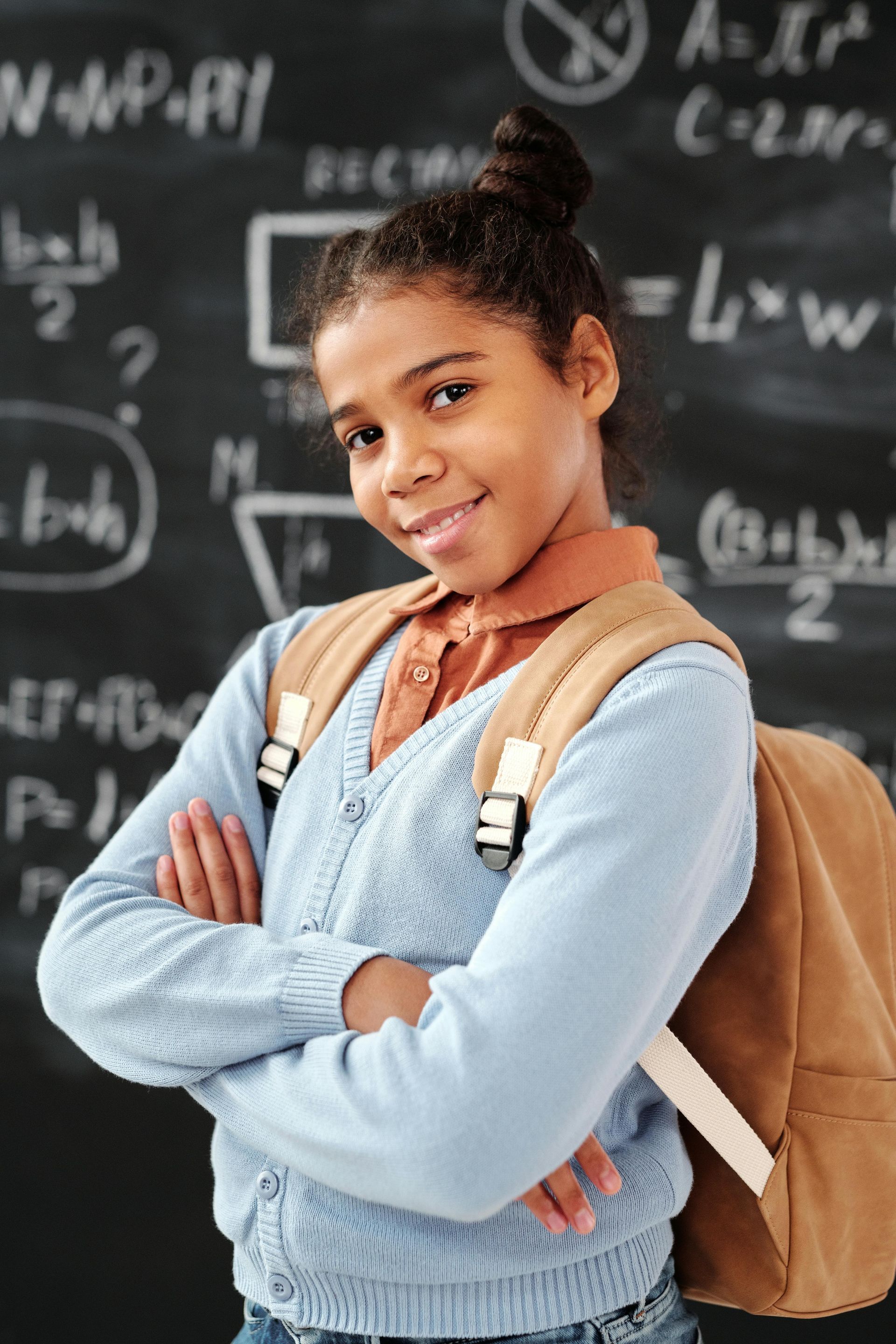 A young girl with a backpack is standing in front of a blackboard.