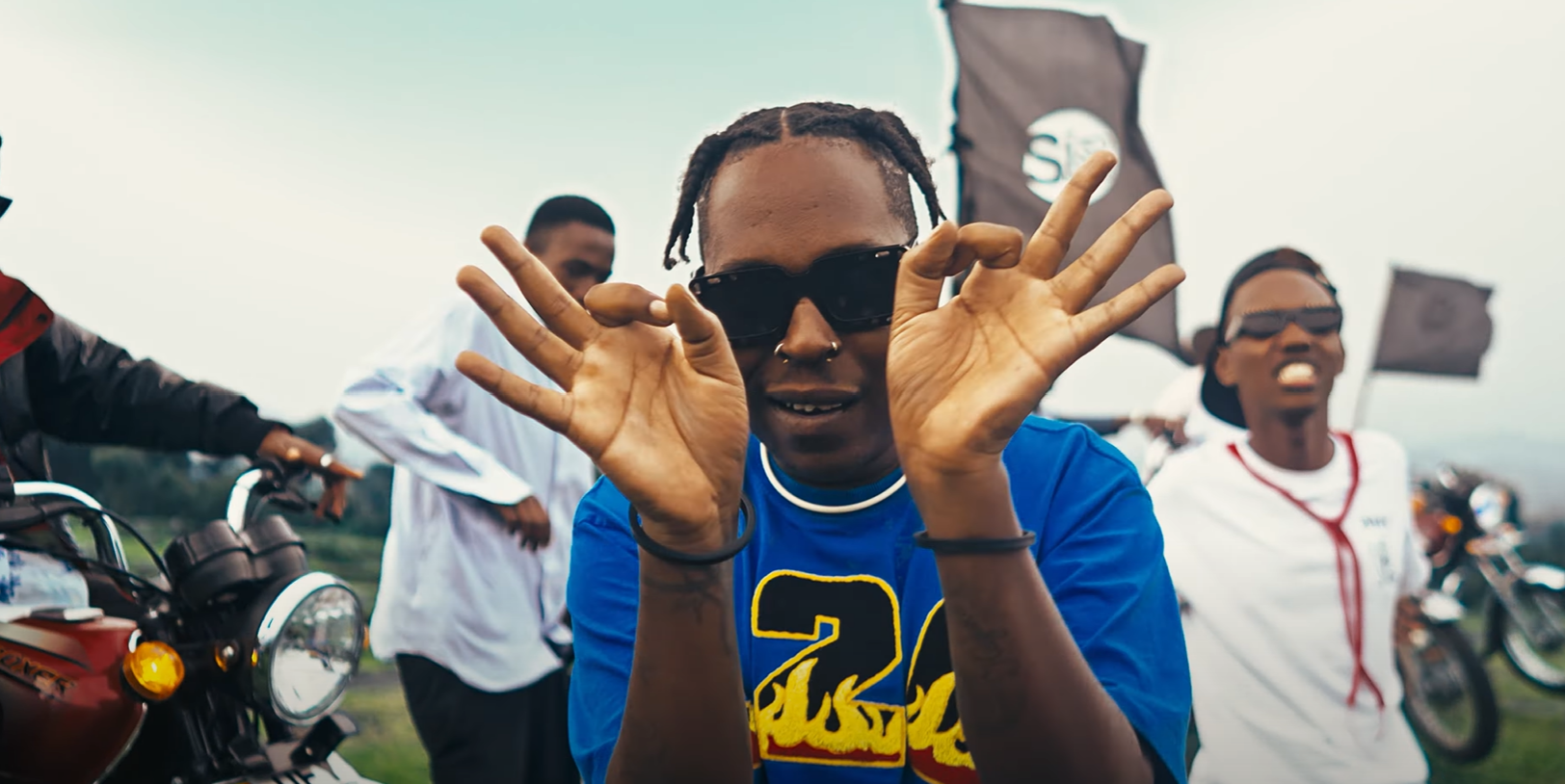 A man wearing sunglasses and a blue shirt is making a peace sign in front of a group of people on motorcycles.