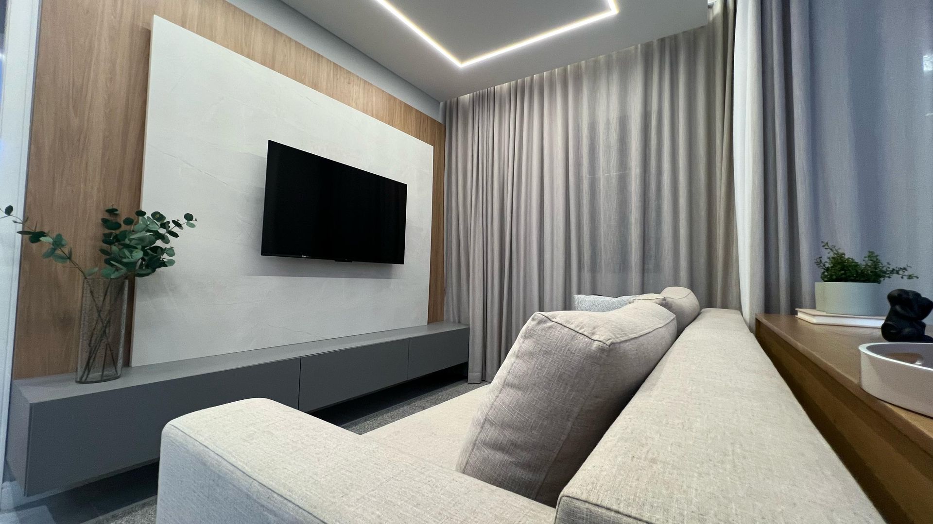 Sala de estar com sofá claro, TV, armário cinza, parede com painéis de madeira e cortinas.