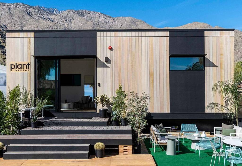 Casa moderna com revestimento de madeira, detalhes em preto, porta de vidro aberta, degraus e área de estar externa. Montanhas ao fundo.