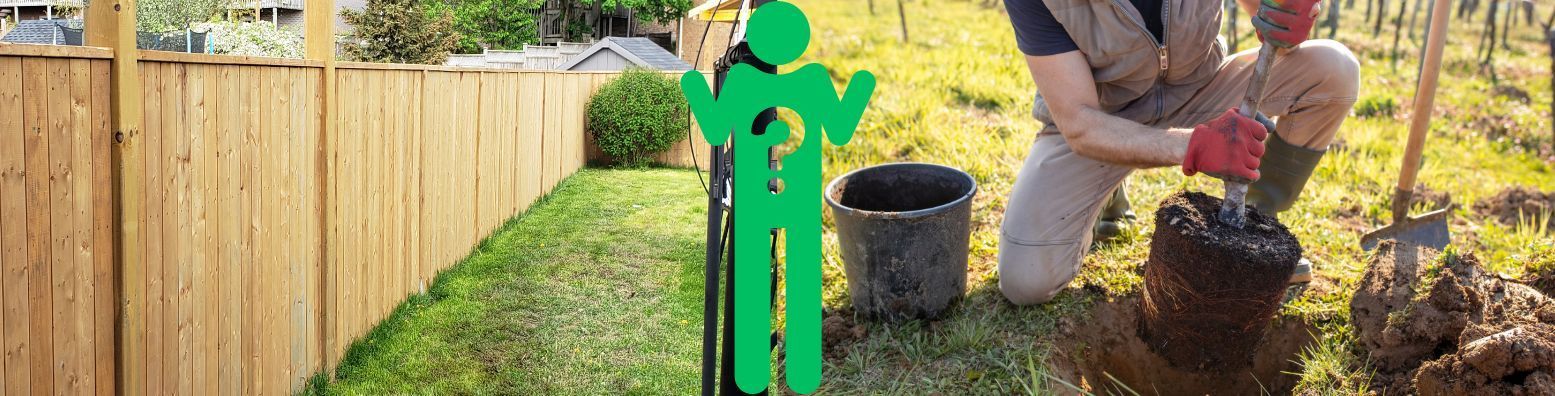 A man is digging a hole in the ground in front of a wooden fence.