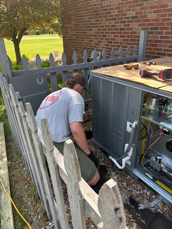 A man is sitting on a fence working on an air conditioner.