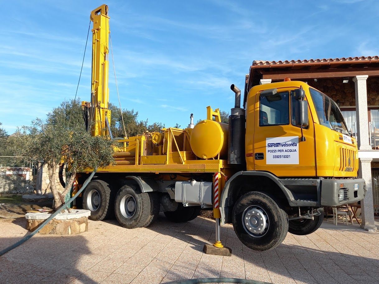 Un camion giallo con una piattaforma di perforazione parcheggiato vicino a un edificio sotto un cielo azzurro, sta perforando un pozzo Sarda Trivelsonda Group Olbia  Loiri Porto San Paolo - Loiri (SS)